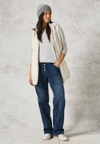 Witte fleecevest over een grijze langemouw T-shirt, gecombineerd met donkerblauwe jeans. Een grijze gebreide beanie en witte sneakers maken de look compleet.