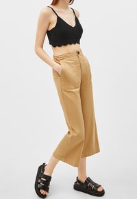 Černý háčkovaný crop top, béžové kalhoty s širokými nohavicemi a předními kapsami, a černé sandály na šněrování. Oblečení má ležérní, uvolněný střih.