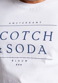 Hvid bomulds t-shirt med blå tekst, der læser "COTCH & SODA," med vandrette linjer og "AMSTERDAMS" og "BLAUW" nedenunder.