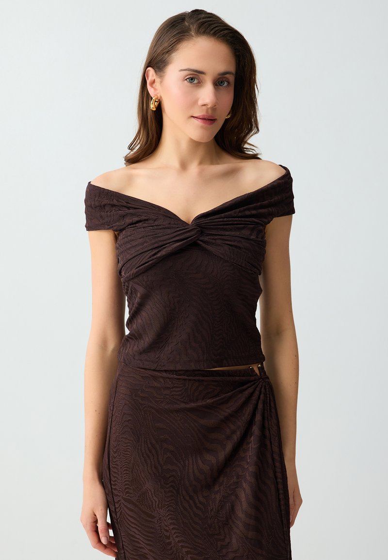 Femme aux cheveux châtain mi-longs portant un top texturé marron foncé épaules dénudées et une jupe assortie, avec des boucles d'oreilles anneaux dorés.