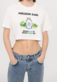 Vit kortärmad t-shirt med en grafik av en burk och texten "Andas in kärlek, andas ut fred" i tjocka svarta och färgglada bokstäver.