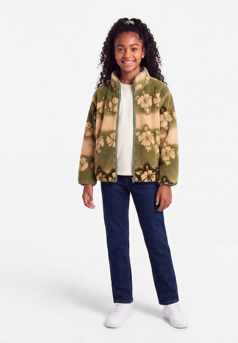 Jeune fille aux cheveux bouclés portant une veste polaire zippée à fleurs vertes et beiges, une chemise blanche, un jean foncé et des baskets blanches, debout et souriante.