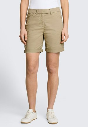 Persoon draagt beige shorts met opgerolde zoom, witte sneakers en een wit topje tegen een effen lichte achtergrond.