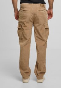 Karamellfarbene Corduroy-Cargo-Hosen mit Seitentaschen, Gesäßtaschen und einem Etikett am Bund. Der Stoff hat eine gerippte Textur.