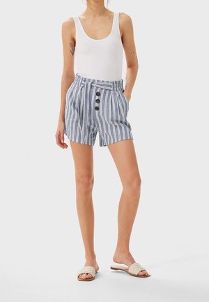 Shorts rayés en tissu bleu et blanc avec un lien à l'avant, quatre boutons et des poches latérales. Portés avec un débardeur blanc et des sandales beiges.