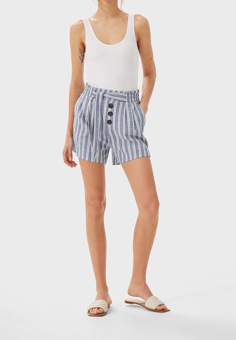 Shorts rayés en tissu bleu et blanc avec un lien à l'avant, quatre boutons et des poches latérales. Portés avec un débardeur blanc et des sandales beiges.