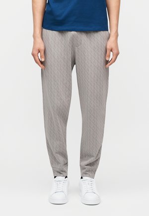 Mężczyzna w jasnoszarych spodniach z subtelnym wzorem, białych trampkach oraz ciemnoniebieskiej koszuli stoi na jednolitym tle.