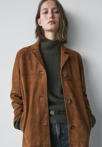Bruine suède jas met een losse pasvorm, voorzien van een reverskraag, drie knopen en twee steekzakken aan de voorkant. Draagt over een donkere geribde coltrui.