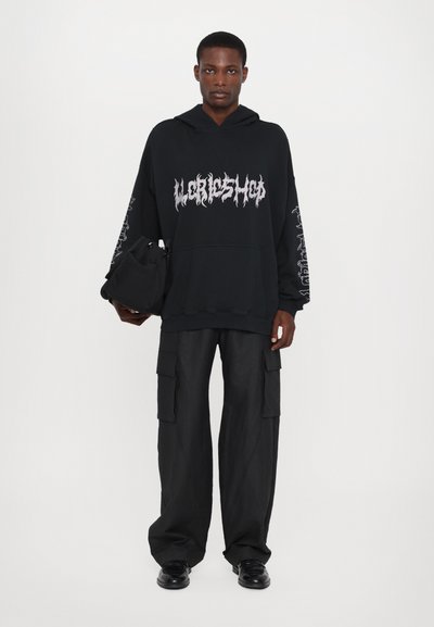 Black oversized hoodie with graphic print, long sleeves, and front pocket; paired with wide-leg black cargo pants and black shoes.