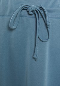 Tessuto di cotone blu con chiusura a cordoncino annodato. La trama è liscia con un leggero lucido. Il dettaglio include un'estremità annodata sul cordoncino.