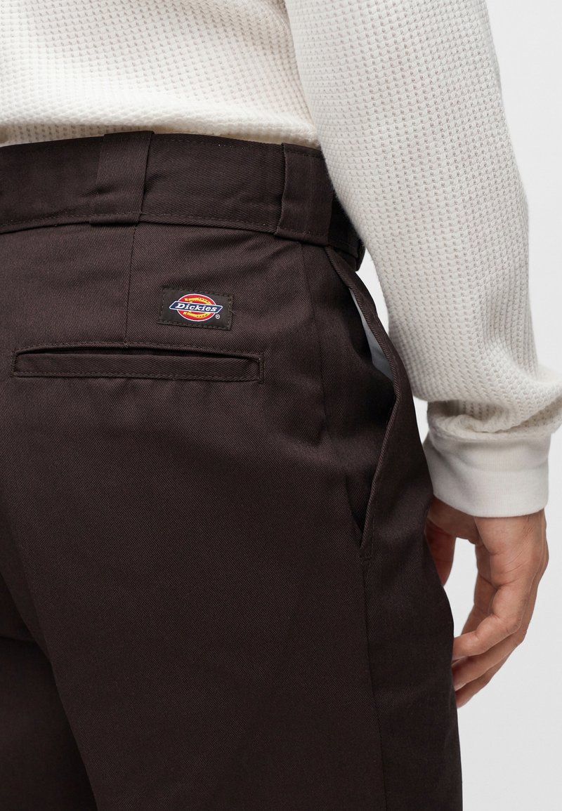 Persona che indossa pantaloni da lavoro marrone scuro con una tasca posteriore e un'etichetta del marchio Dickies, abbinati a una camicia bianca testurizzata a maniche lunghe.