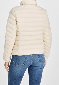Osoba ubrana w kremową pikowaną kurtkę puchową i niebieskie dżinsy, pokazana z tyłu na jednolitym tle.