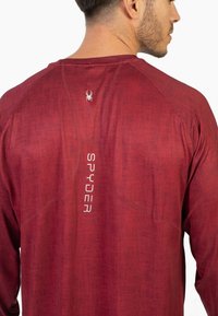 Punainen pitkähihainen paita, jossa teksturoitu kangas, taakse ommeltu valkoinen "SPYDER" -logo ja hämähäkkisymboli, muotoiltu istuvuus raglan-hihalla.