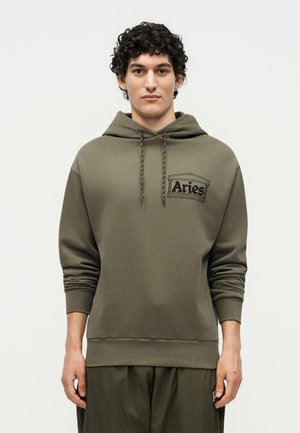 Junger Mann mit lockigem Haar, der einen olivgrünen Hoodie mit dem „Aries“-Logo auf der Brust trägt und vor einem schlichten hellen Hintergrund steht.