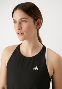 Débardeur de sport noir en tissu texturé, avec un logo Adidas blanc sur la poitrine et de fines bretelles blanches en dessous.