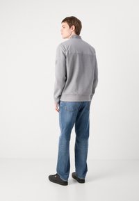 Pulover gri cu guler înalt și spate uniform, asortat cu blugi denim albaștri. Modelul poartă pantofi negri și are părul scurt, de culoare brunetă.