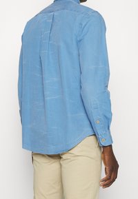 Blue long-sleeve shirt with a smooth texture, buttoned cuffs, back pleat, and curved hem, displayed on a model in beige pants.