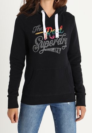Sweat à capuche noir avec cordons blancs et texte coloré "The Real Superdry JPN" porté avec un jean bleu.