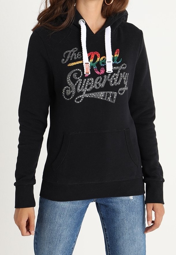 Sweat à capuche noir avec cordons blancs et texte coloré "The Real Superdry JPN" porté avec un jean bleu.