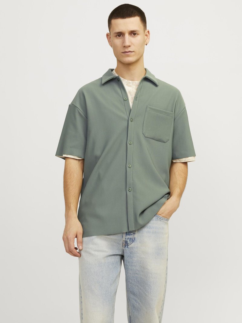 Camicia a maniche corte con bottoni in morbido tessuto verde, dotata di tasca, con una sottile trama e una vestibilità rilassata, abbinata a jeans blu chiaro.