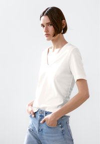 Biała koszulka z krótkim rękawem w serek, wykonana z gładkiego materiału, zestawiona z jasnoniebieskimi dżinsami. Prosty design, luźny krój, minimalistyczny wygl�ąd.