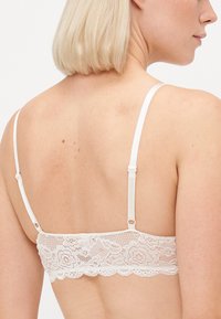 Witte kant bralette met delicate bloemenpatronen, verstelbare bandjes en een haak-en-oogsluiting aan de achterkant. Zachte, gestructureerde stof.