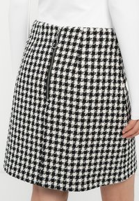 Czarno-biała spódnica w wzór houndstooth z zamkiem błyskawicznym z przodu, noszona z białą bluzką z długim rękawem, pokazująca prawą dłoń spoczywającą blisko dołu.