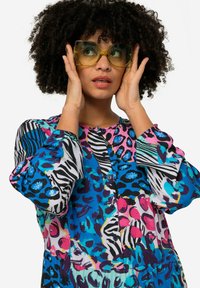 Blouse colorée à motifs présentant des imprimés animaliers vibrants en bleu, rose et noir, avec un décolleté rond et des détails de boutons ; manches longues.