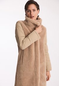 Długi, beżowy płaszcz z puszystą, teksturowaną częścią przednią i gładkimi, pikowanymi rękawami. Wysoki kołnierz i zapięcie z przodu.