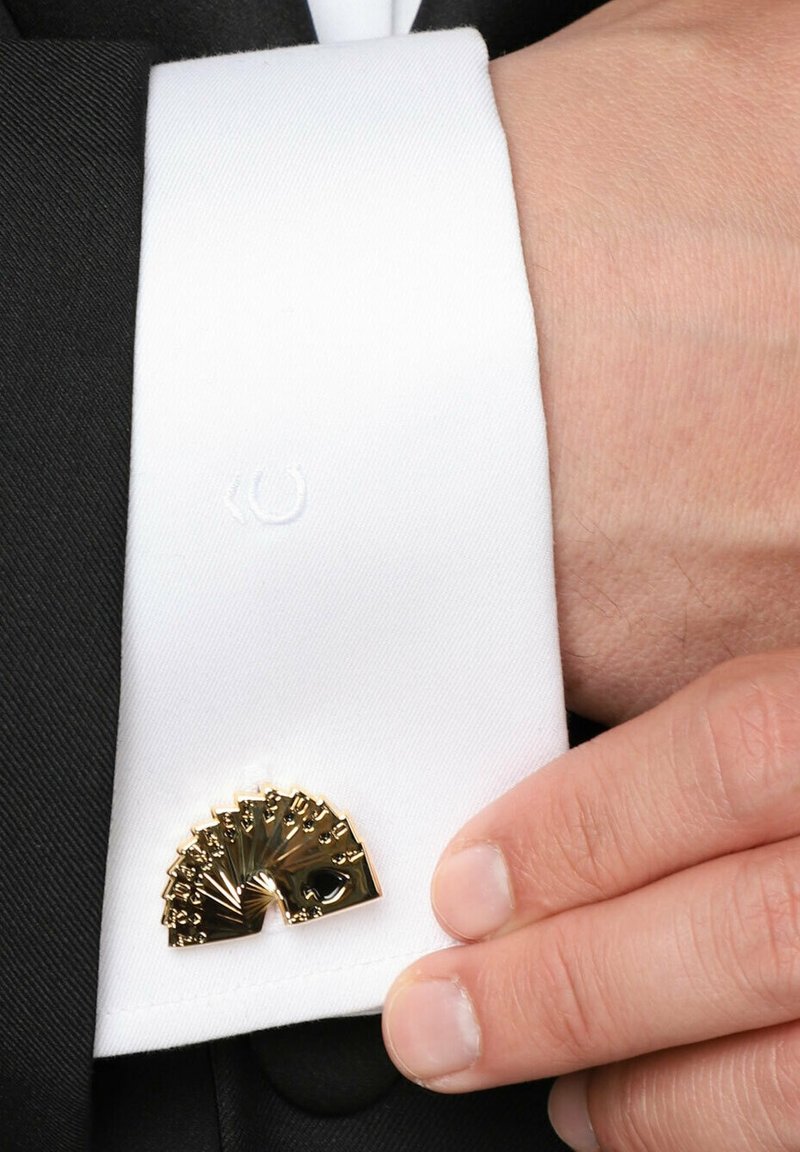 Złote spinka do mankietów w kształcie wachlarza na białym mankiecie, z detalicznymi akcentami. Widoczna czarna tkanina garnituru, podkreślająca kontrast mankietu.