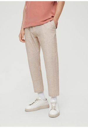 Uomo che indossa pantaloni beige alla caviglia, calzini bianchi, sneakers bianche e una maglietta color corallo con una mano in tasca.