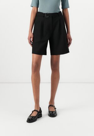 Personne portant un short noir ceinturé aux genoux, un haut bleu clair et des chaussures Mary Jane en cuir verni noir, debout devant un fond blanc.