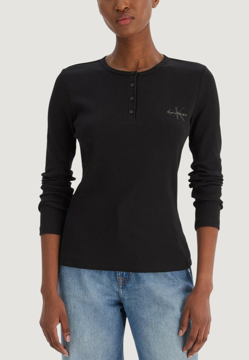 Czarna, prążkowana koszulka z długim rękawem, z dekoltem henley, trzema guzikami z przodu oraz małym logo na piersi. Połączona z jasnoniebieskimi dżinsami.