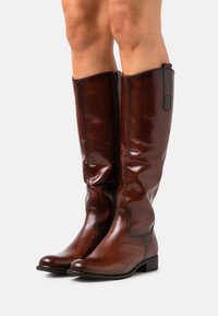 Bottes hautes en cuir marron brillant, zippées sur le côté intérieur, portées sur des jambes nues sur fond blanc uni.