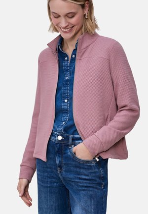 Femme souriante portant une veste zippée rose poussiéreuse texturée par-dessus une chemise en jean bleue et un jean, une main dans la poche et la tête légèrement inclinée vers le bas.