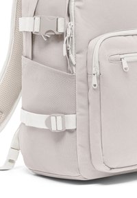 Sac à dos gris clair en tissu texturé, avec plusieurs zips, des bretelles réglables et une poche avant avec fermeture éclair.