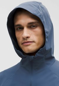 Blauwe waterdichte jas met een hoge kraag en capuchon, gemaakt van soepel materiaal. Beschikt over een ritssluiting aan de voorkant en minimale naden voor een slanke uitstraling.