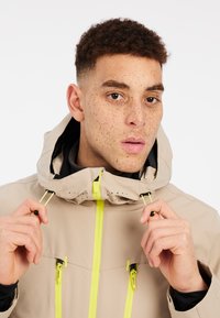 Beige Überjacke mit Kapuze, akzentuiert mit leuchtend gelben Reißverschlüssen und Beschlägen, mit einer glatten Textur und Belüftungslöchern.