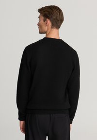 Czarny sweter robiony na drutach z okrągłym dekoltem, żebrowymi mankietami i dołem, o miękkiej fakturze i luźnym kroju. Pokazany widok z tyłu.