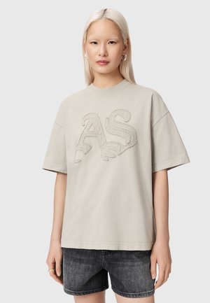 Jeune femme aux cheveux blonds raides portant un T-shirt beige surdimensionné avec les lettres "AS" en relief et un short en jean noir, posant devant un fond uni.