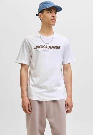 Balta trumpa rankovėmis marškinėliai su tamsiai rudu "JACK&JONES" logotipo spauda ir mažu piešinėliu. Dėvimi su šviesiai pilkais sportiniais kelniais ir mėlynu kepurėle.