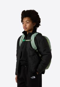 Green backpack with padded straps and a buckle, worn over a black quilted jacket and black pants with a logo, presenting a casual, layered look.