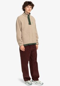 Beige fleece pullover with a half-zip, green collar accent, and textured fabric. Paired with dark red corduroy pants and running shoes.