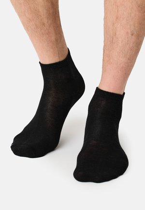 Männliche Beine mit niedrig geschnittenen schwarzen Knöchelsocken, die auf einer weißen Oberfläche stehen.