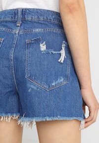 Pantalones cortos de mezclilla azul con dobladillo deshilachado, acentos desgastados y un diseño de cintura alta. Presenta bolsillos con un detalle rasgado en el lado derecho.