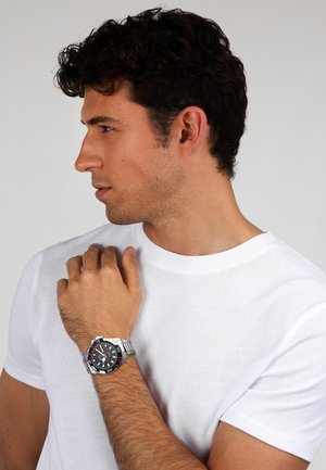Orologio sportivo argentato con quadrante nero, indici luminescenti e bracciale in acciaio inossidabile esposto su una mano. Il modello indossa una t-shirt bianca.