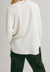 White long-sleeve blouse with a smooth texture, featuring buttoned cuffs and a relaxed fit, paired with dark green pants.