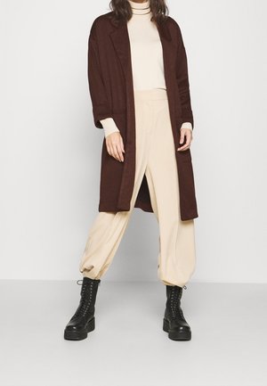 Vrouw die een beige coltrui en broek draagt, een donkerbruine lange jas en zwarte veterplatformlaarzen, staand op een grijze vloer.