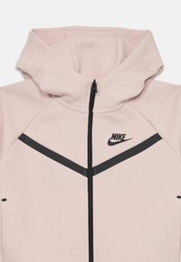 Moletom rosa claro com fecho zip e um logotipo e detalhes em preto. Apresenta capuz, corte ajustado e riscas diagonais pretas em contraste na parte da frente.
