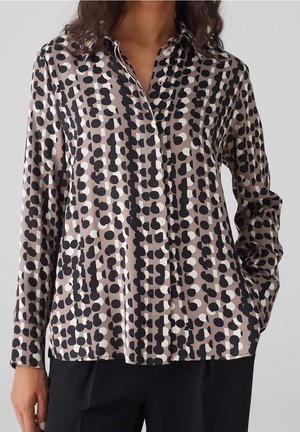 Blouse à boutons en tissu soyeux. Présente un fond beige avec des motifs circulaires noirs et blancs. Manches longues et col détaillé.